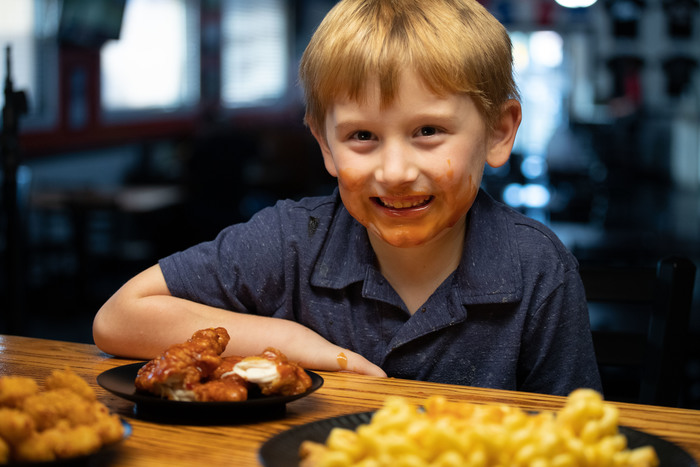 A young guest at The Dirty Buffalo Little Creek grinning with buffalo sauce on his face beside a plate of wings