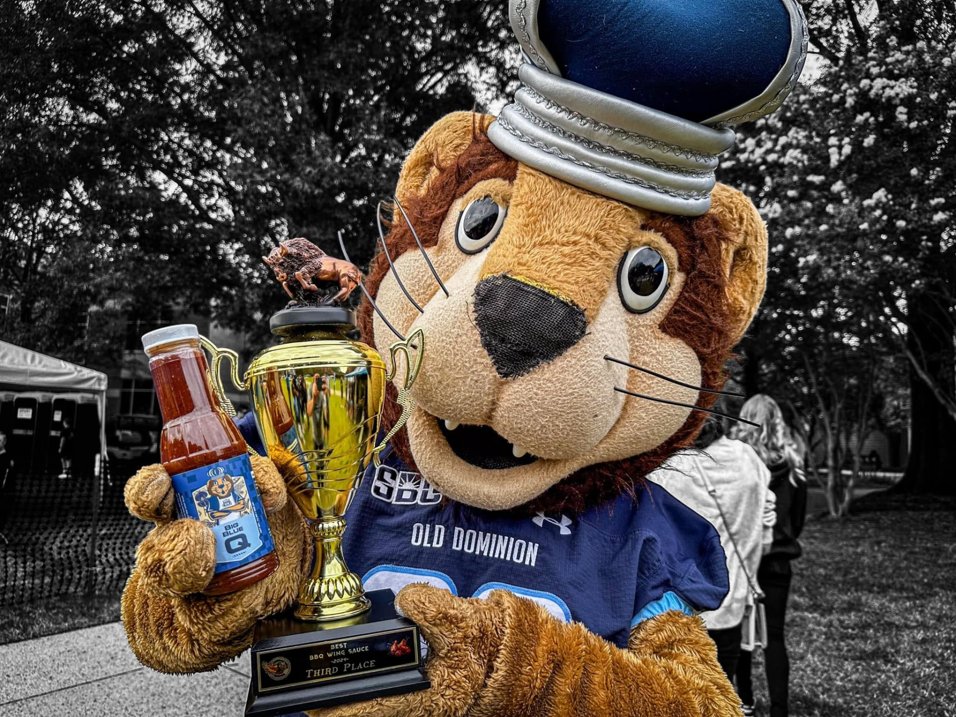ODU mascot Big Blue holding a bottle of Big Blue Q sauce and the Best BBQ Wing Sauce trophy